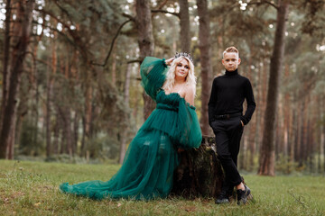 A beautiful blonde young woman in a long green dress and a diadem on her head with stylish young son in the forest. girl and boy sitting near the old stump. Solar glare. Fantasy. fairy tale.