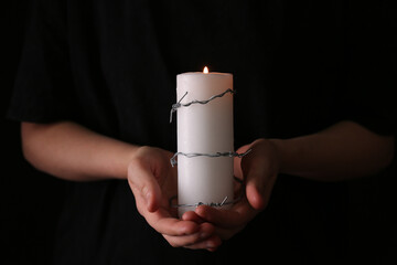 Woman holding candle with barbed wire on black background, closeup. Holocaust memory day