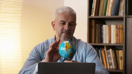 Mature man looking at globe map of world and dreaming of vacation, sitting near laptop at home - Powered by Adobe