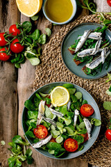 Green field salad with pickled anchovies or sardines fillet, and cherry tomatoes, served in blue bowl with lemon and olive oil on straw napkin over old wooden background. Flat lay, space