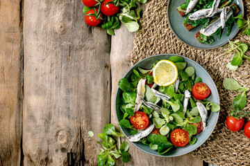 Green field salad with pickled anchovies or sardines fillet, and cherry tomatoes, served in blue bowl with lemon and olive oil on straw napkin over old wooden background. Flat lay, space