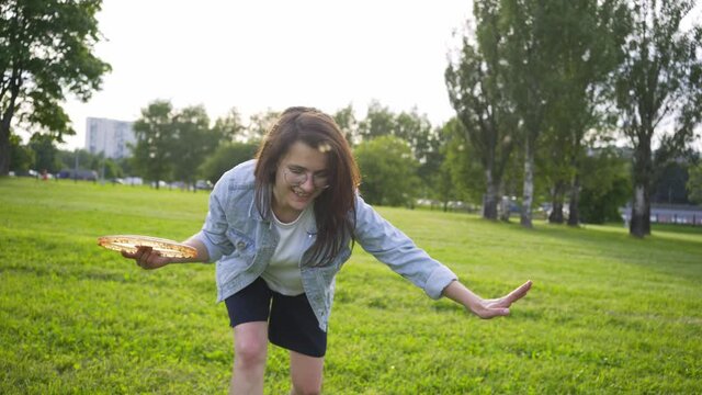 Close-up portrait cheerful brunette woman in denim jacket and glasses playing frisbee with pet. Lady throwing plate while training tricks with funny large watchdog on fresh green grass in city park 