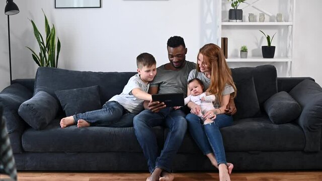 A multiracial family relaxing with a digital tablet sitting on the comfortable sofa, watching video, playing game on the touchscreen, have fun together. Spouses, baby girl and school-age son