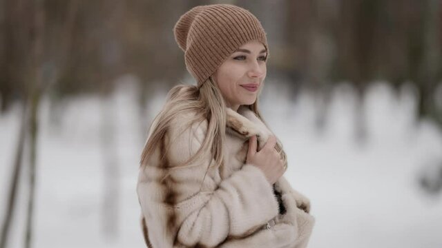 Portrait of a fashionable blonde in a beautiful fur coat and a beige hat posing and smiling against the background of a winter park. The model is located on the street on a snowy background.