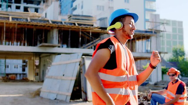 Afro American worker at construction site dancing charismatic while have a break listening music from the headphones he wearing safety equipment at work