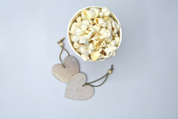 Selective focus top view of small light beige wooden necklaces in form of heart and appetizing popcorn in round paper disposable cup in light of lamps on white background 