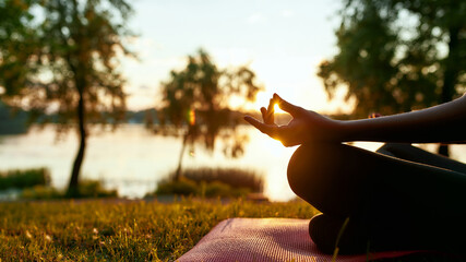Cropped shot of a woman doing yoga outdoors near lake or river at sunrise in the morning, sitting in lotus pose on the background of nature