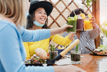 Young multiracial women enjoy brunch together and cheering with smoothies while wearing surgical...