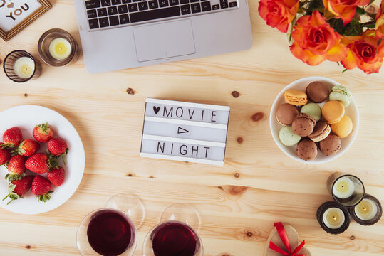 Top view Romantic Movie night concept. Movie night message on light board, laptop, candles, flowers, macaroons, berries, and two glasses with wine for belovers. Cozy holiday plans for Valentine's day.