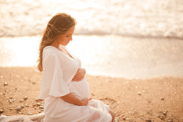 Pregnant woman wearing white dress sittin over sea shore outdoors. Motherhood. Maternity. Healthy lifestyle.