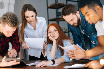 Fototapeta premium Exhausted tired young business woman sitting at desk, colleagues shaking papers in face paper documents. Businesswoman under stress at workplace. Female boss exhausted by management work.