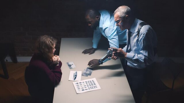 Diverse detective interrogating scared arrested woman and threatening with gun