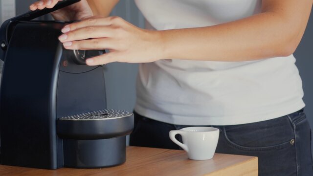A mixed-race woman in a white t-shirt putting a compostable coffee capsule into the coffee machine. White ceramic cup on the table. High-resolution jpg photo
