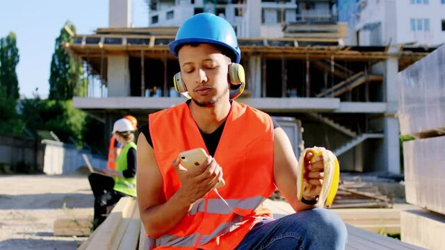 Young construction worker Afro American at the break time eating a banana and listening music in front of the camera at construction site