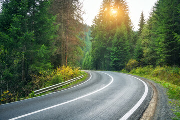 Road in forest at sunset in spring. Beautiful mountain roadway, trees with green and orange foliage. Landscape with empty asphalt winding road through woodland in autumn. Travel in Europe. Nature