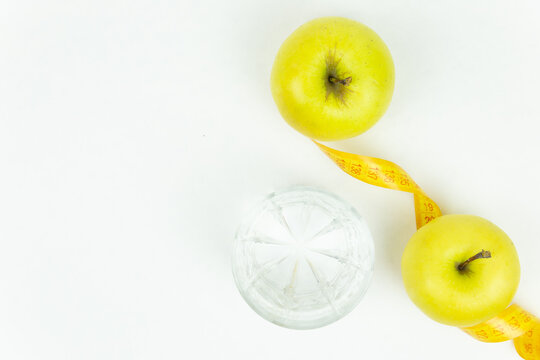 A glass of water and a measuring tape wrapped around two green apples as a symbol of diet. The concept of a healthy lifestyle, food and sports, on a white background. Top view with place for text or