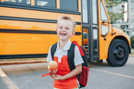 Happy laughing smiling Caucasian boy student by yellow bus on first September day. Hard of hearing child kid eating apple fruit at school yard outdoors. Education and back to school in Autumn Fall.