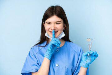 Young dentist woman holding tools over isolated blue background doing silence gesture