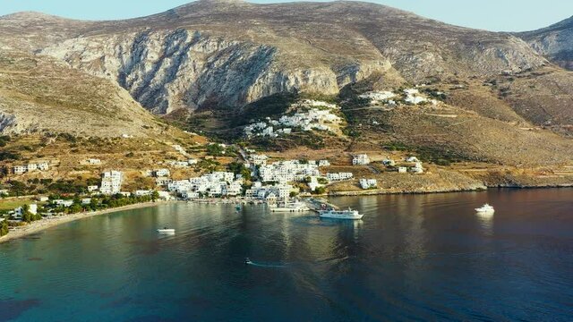 Drone video of Aegialis in Greece. Drone flies high over the small town, that is surrounded by mountains, and clear blue water. There is a small marina, and there is a boat sailing towards the harbour
