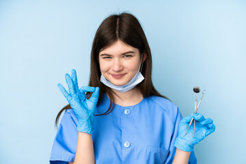 Young dentist woman holding tools over isolated blue background showing an ok sign with fingers