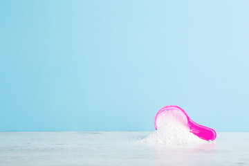 Pink cup or scoop of white powder on table. Detergent for clothes washing. Empty place for text or logo on light blue wall background. Pastel color. Front view. Closeup.