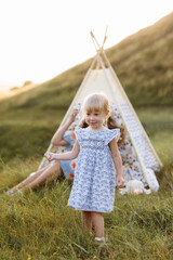 Beautiful mother with her little daughter sitting near wigwam in the field. Spending time together, outside, on vacation, outdoors. Beautiful sunset light in the garden or in the park. © Andriy Medvediuk