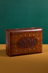 An old vintage handcarved wooden box in a dark brown color with golden details on a yellow surface with a green background. An antique authentic handmade woodcarved treasure container
