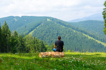 Fototapeta na wymiar Guy sitting on log and enjoy peaceful green mountains landscape. Peace of mind and relax