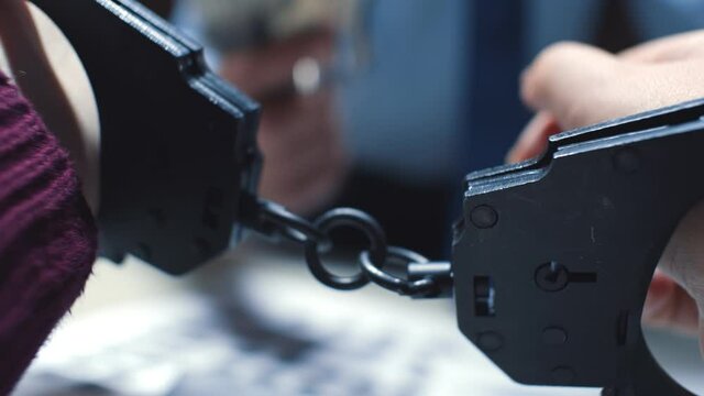 Close up of arrested criminal hands in handcuffs with detective interrogating on background