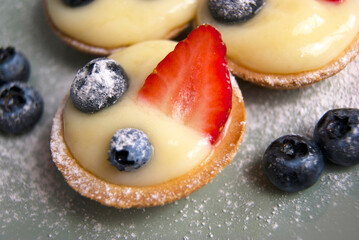 Close up of French desserts. Little tarts with custard, fresh berries and powdered sugar. Tartlets with strawberries, blueberries served with icing sugar (castor sugar) on gray and white plates
