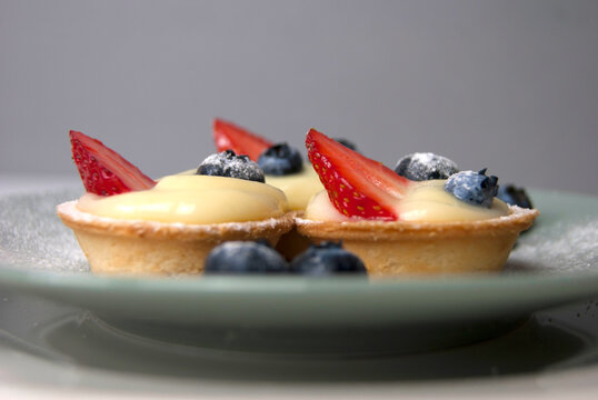 Side view of French dessert. Little tarts with custard, fresh berries and powdered sugar. Tartlets with strawberries, blueberries served with icing sugar (castor sugar) on gray and white plates
