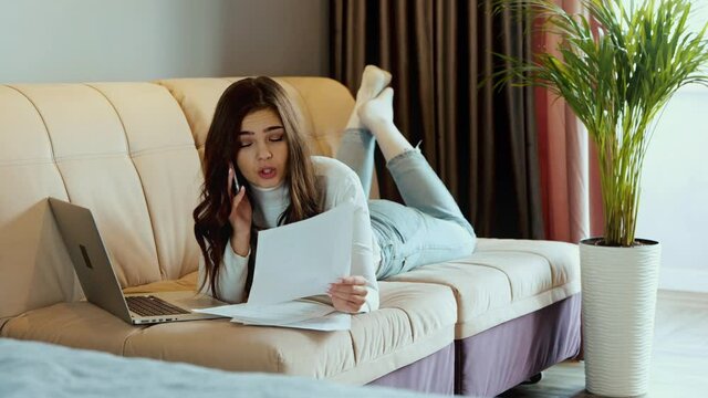 Young brunette woman is lying on the sofa and using smartphone, laptop, documents. The female model has a telephone conversation. Distance education or work from home.