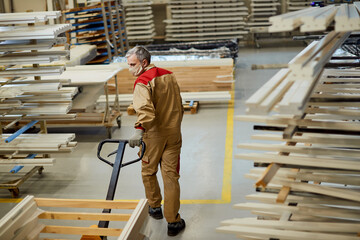 Fototapeta premium Full length of carpenter with face mask pulling pallet jack in a workshop.