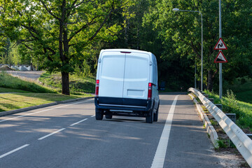 White cargo delivery van driving on a downhill countryside road in summer under green trees
