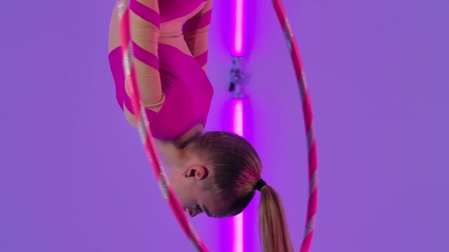 Aerial acrobat in the ring. A young girl in a pink leotard performs acrobatic elements on the air hoop. Shot in the studio with bright neon lights in the background. Close up. Slow motion.