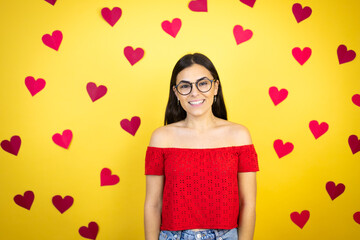 Young beautiful woman over yellow background with red hearts with a happy face standing and smiling with a confident smile showing teeth