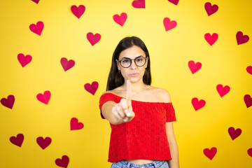 Obraz na płótnie Canvas Young beautiful woman over yellow background with red hearts showing and pointing up with fingers number one while is serious