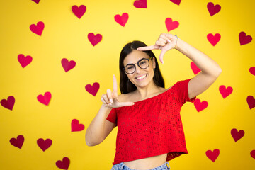 Obraz na płótnie Canvas Young beautiful woman over yellow background with red hearts smiling making frame with hands and fingers with happy face