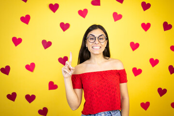 Obraz na płótnie Canvas Young beautiful woman over yellow background with red hearts showing and pointing up with fingers number one while smiling confident and happy