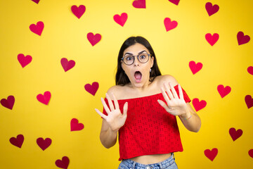 Young beautiful woman over yellow background with red hearts afraid and terrified with fear expression stop gesture with hands, shouting in shock. Panic concept.