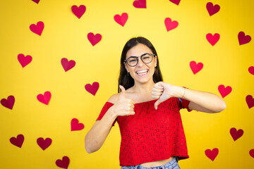 Young beautiful woman over yellow background with red hearts Doing thumbs up and down, disagreement and agreement expression. Crazy conflict