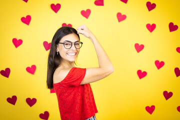 Young beautiful woman over yellow background with red hearts feeling happy, satisfied and powerful, flexing fit and muscular biceps, looking strong after the gym