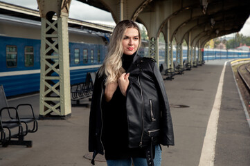 Fototapeta premium Portrait of a young beautiful woman in a leather jacket on a perone next to the train at the railway station. Waiting for the train Vitebsk station St. Petersburg.