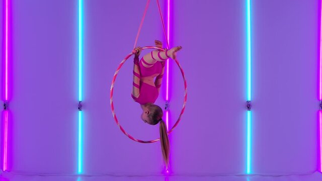 Aerial acrobat in the ring. A young girl in a pink leotard performs acrobatic elements on the air hoop. Shot in the studio with bright neon lights in the background. Slow motion.