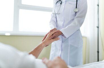 Confident doctor in a white coat holds a hand of a patient while he is lying on the bed in the hospital ward