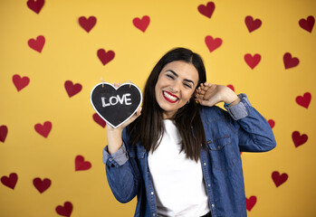 Young caucasian woman over yellow background with red hearts holding heart a blackboard with love word message celebrating crazy and amazed for success with open eyes screaming excited. winner concept