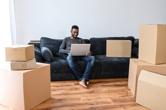 An African-American guy chooses new furniture online in his new house sitting on the couch in empty living room among cardboard boxes, a multiracial man with laptop on the sofa at moving day