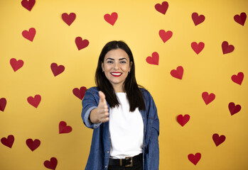 Young caucasian woman over yellow background with red hearts smiling friendly offering handshake as greeting and welcoming