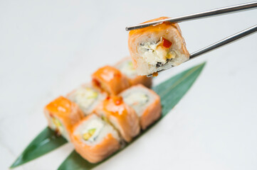 Close-up of Sushi Roll piece with salmon Tataki on top hold in metal chopsticks with blurred Japanese dish with avocado and Philadelphia cream cheese served with Thai sauce on white marble background
