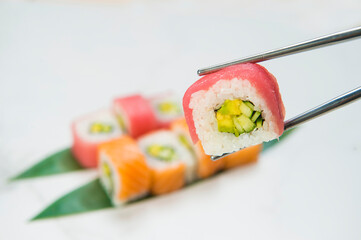 Close up of Sushi Roll piece with salmon and tuna slices on top hold in metal chopsticks with blurred Japanese dish with avocado and cucumber inside on white marble background
